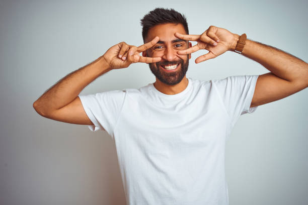 istock Young indian man wearing t-shirt standing over isolated white background Doing peace symbol with fingers over face, smiling cheerful showing victory 1205736631