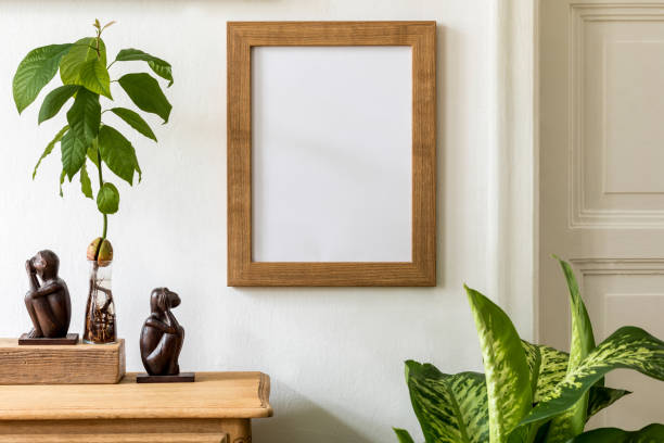 Minimalistic compositon with wooden vintage commode, brown mock up photo frame, avocado plant, plants and elegant personal accessories. Stylish living room. White walls. Template.