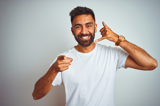 Young indian man wearing t-shirt standing over isolated white background smiling doing talking on the telephone gesture and pointing to you. Call me.