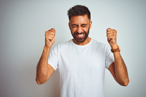 Young indian man wearing t-shirt standing over isolated white background excited for success with arms raised and eyes closed celebrating victory smiling. Winner concept.