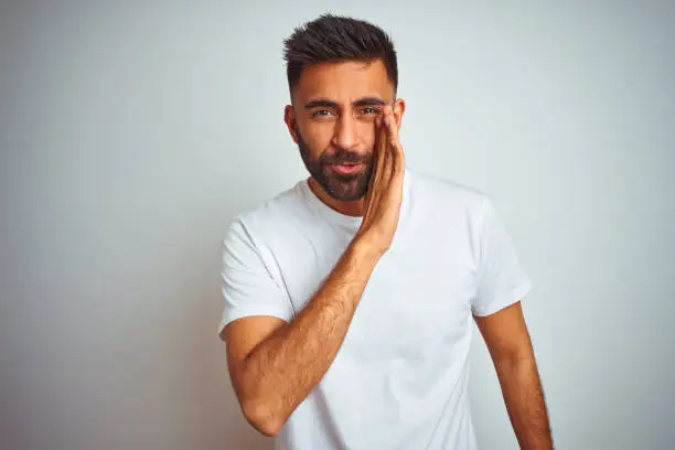 Photo of Young indian man wearing t-shirt standing over isolated white background hand on mouth telling secret rumor, whispering malicious talk conversation