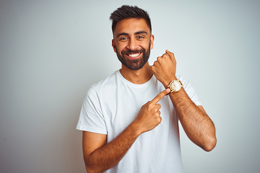 Young indian man wearing t-shirt standing over isolated white background In hurry pointing to watch time, impatience, looking at the camera with relaxed expression
