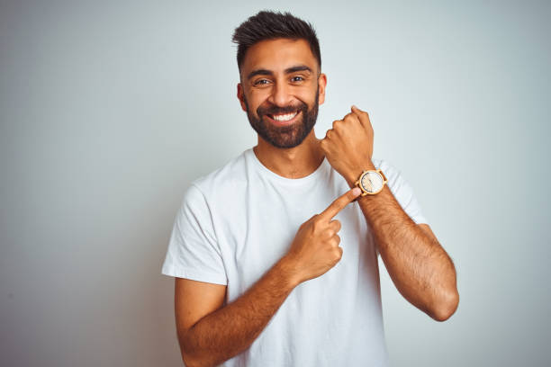 young indian man wearing t-shirt standing over isolated white background in hurry pointing to watch time, impatience, looking at the camera with relaxed expression - clock face fotos imagens e fotografias de stock