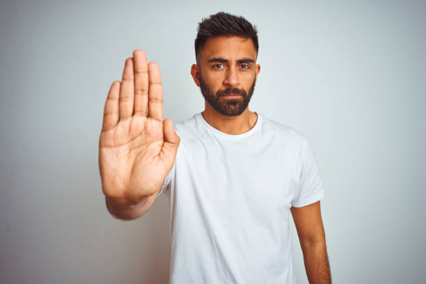 istock Young indian man wearing t-shirt standing over isolated white background doing stop sing with palm of the hand. Warning expression with negative and serious gesture on the face. 1205737149