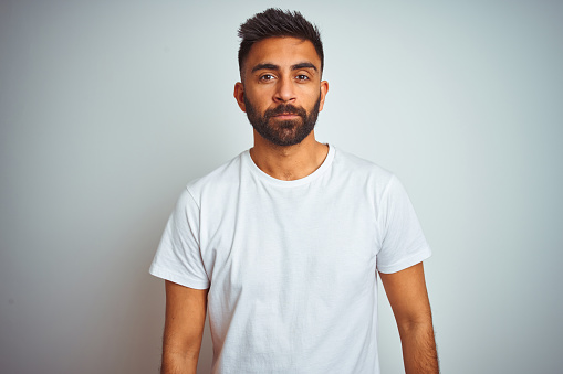 Young indian man wearing t-shirt standing over isolated white background Relaxed with serious expression on face. Simple and natural looking at the camera.