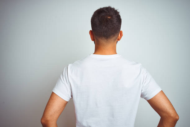 молодой индийский человек в футболке, стоящей над изолированным белым фоном, стоящим назад, глядя в сторону с руками на теле - назад стоковые фото и изображения