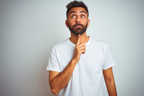 istock Young indian man wearing t-shirt standing over isolated white background Thinking concentrated about doubt with finger on chin and looking up wondering 1205738537
