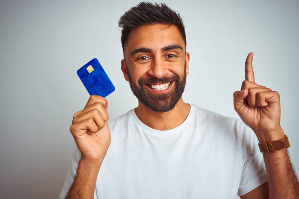 junge indische kunden mann hält kreditkarte stehen über isolierten weißen hintergrund überrascht mit einer idee oder frage zeige finger mit glücklichem gesicht, nummer eins - surprise ideas business person pointing stock-fotos und bilder