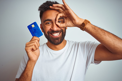 Young indian customer man holding credit card standing over isolated white background with happy face smiling doing ok sign with hand on eye looking through fingers