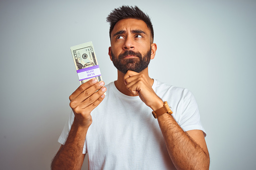 Young indian man holding dollars standing over isolated white background serious face thinking about question, very confused idea