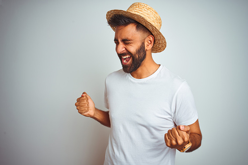 Young indian man on holiday wearing summer hat standing over isolated white background very happy and excited doing winner gesture with arms raised, smiling and screaming for success. Celebration concept.