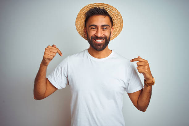 Young indian man on holiday wearing summer hat standing over isolated white background looking confident with smile on face, pointing oneself with fingers proud and happy.