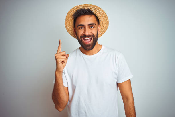 istock Young indian man on holiday wearing summer hat standing over isolated white background pointing finger up with successful idea. Exited and happy. Number one. 1205739267