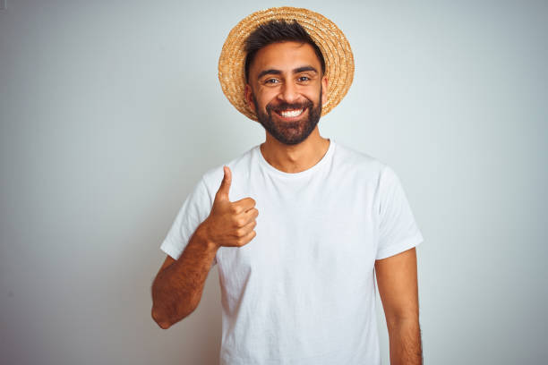 istock Young indian man on holiday wearing summer hat standing over isolated white background doing happy thumbs up gesture with hand. Approving expression looking at the camera with showing success. 1205739382