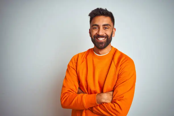 Photo of Young indian man wearing orange sweater over isolated white background happy face smiling with crossed arms looking at the camera. Positive person.