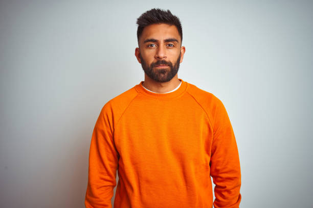 istock Young indian man wearing orange sweater over isolated white background Relaxed with serious expression on face. Simple and natural looking at the camera. 1205739665