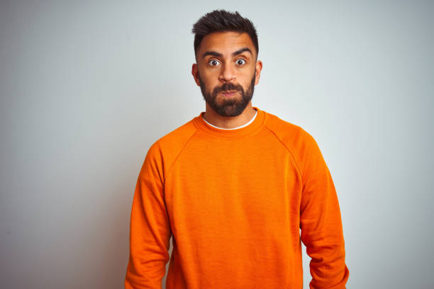 istock Young indian man wearing orange sweater over isolated white background puffing cheeks with funny face. Mouth inflated with air, crazy expression. 1205739695