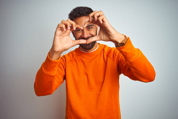 junge indische mann trägt orange pullover über isolierten weißen hintergrund lächelnd in liebe herzsymbol form mit händen. romantisches konzept. - smiling single object photography orange stock-fotos und bilder