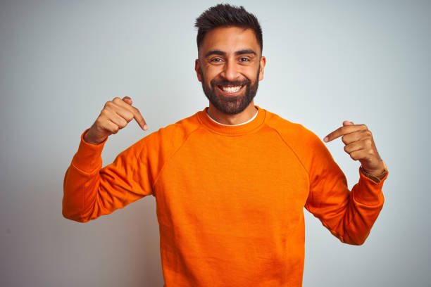 istock Young indian man wearing orange sweater over isolated white background looking confident with smile on face, pointing oneself with fingers proud and happy. 1205739879