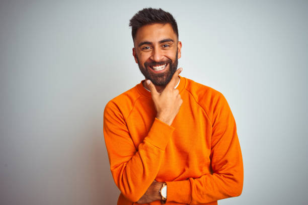 istock Young indian man wearing orange sweater over isolated white background looking confident at the camera smiling with crossed arms and hand raised on chin. Thinking positive. 1205739948