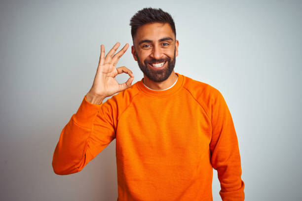 istock Young indian man wearing orange sweater over isolated white background smiling positive doing ok sign with hand and fingers. Successful expression. 1205740006