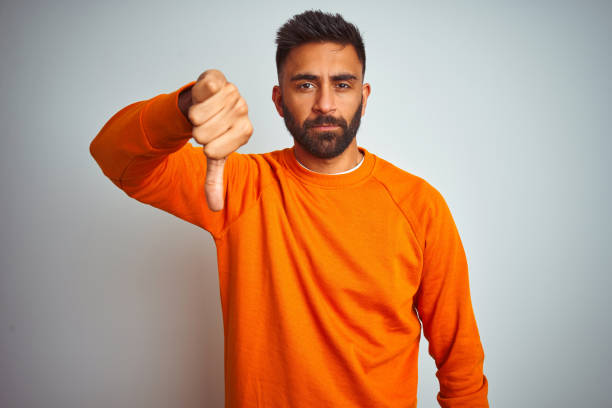 istock Young indian man wearing orange sweater over isolated white background looking unhappy and angry showing rejection and negative with thumbs down gesture. Bad expression. 1205740077