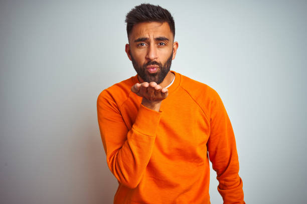istock Young indian man wearing orange sweater over isolated white background looking at the camera blowing a kiss with hand on air being lovely and sexy. Love expression. 1205740603