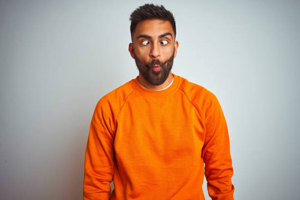 istock Young indian man wearing orange sweater over isolated white background making fish face with lips, crazy and comical gesture. Funny expression. 1205740631