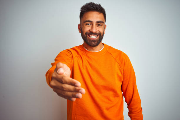 istock Young indian man wearing orange sweater over isolated white background smiling friendly offering handshake as greeting and welcoming. Successful business. 1205740632