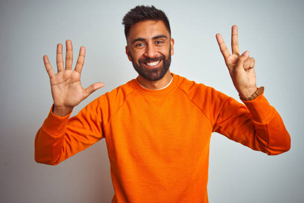 junger indischer mann trägt orangefarbenen pullover über isoliertem weißen hintergrund zeigt und zeigt mit den fingern nummer sieben, während er selbstbewusst und glücklich lächelt. - hispanic male stock-fotos und bilder