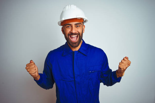 istock Handsome indian worker man wearing uniform and helmet over isolated white background celebrating surprised and amazed for success with arms raised and open eyes. Winner concept. 1205740748
