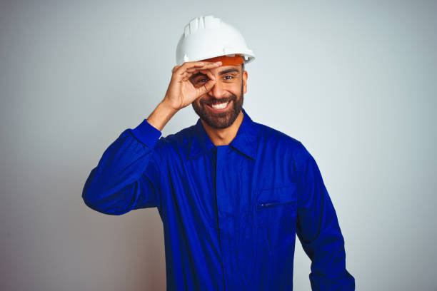 istock Handsome indian worker man wearing uniform and helmet over isolated white background doing ok gesture with hand smiling, eye looking through fingers with happy face. 1205740783