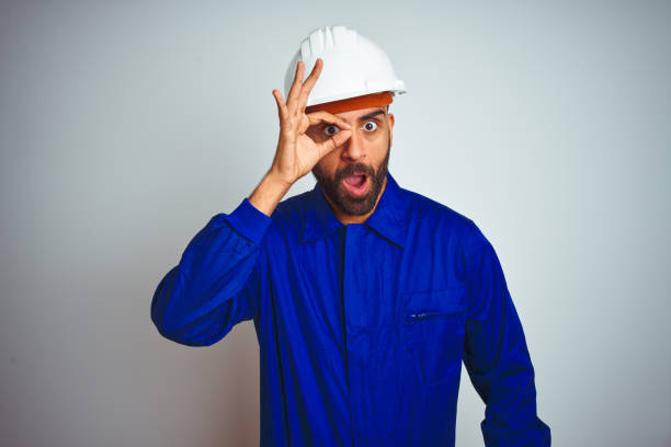 istock Handsome indian worker man wearing uniform and helmet over isolated white background doing ok gesture shocked with surprised face, eye looking through fingers. Unbelieving expression. 1205740784