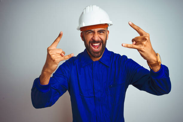 istock Handsome indian worker man wearing uniform and helmet over isolated white background shouting with crazy expression doing rock symbol with hands up. Music star. Heavy concept. 1205740808