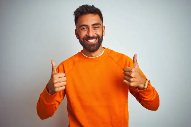 Young indian man wearing orange sweater over isolated white background success sign doing positive gesture with hand, thumbs up smiling and happy. Cheerful expression and winner gesture.