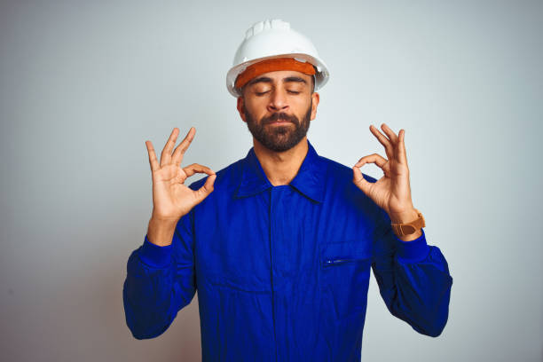Handsome indian worker man wearing uniform and helmet over isolated white background relax and smiling with eyes closed doing meditation gesture with fingers. Yoga concept.