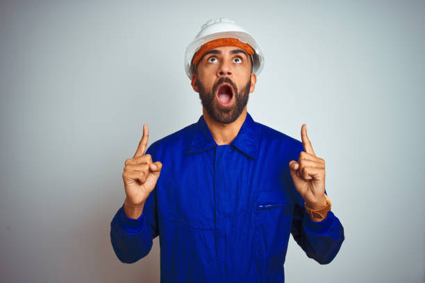 istock Handsome indian worker man wearing uniform and helmet over isolated white background amazed and surprised looking up and pointing with fingers and raised arms. 1205741095