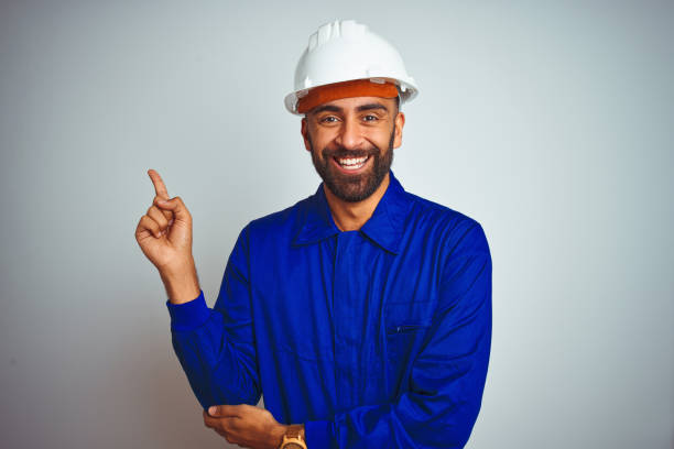 Handsome indian worker man wearing uniform and helmet over isolated white background with a big smile on face, pointing with hand and finger to the side looking at the camera. Handsome indian worker man wearing uniform and helmet over isolated white background with a big smile on face, pointing with hand and finger to the side looking at the camera. sign human hand pointing manual worker stock pictures, royalty-free photos & images