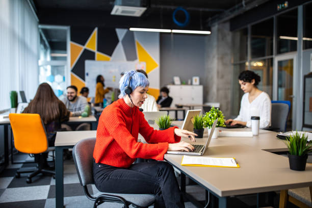 istock Young woman working on laptop with headset in busy modern office 1205741107