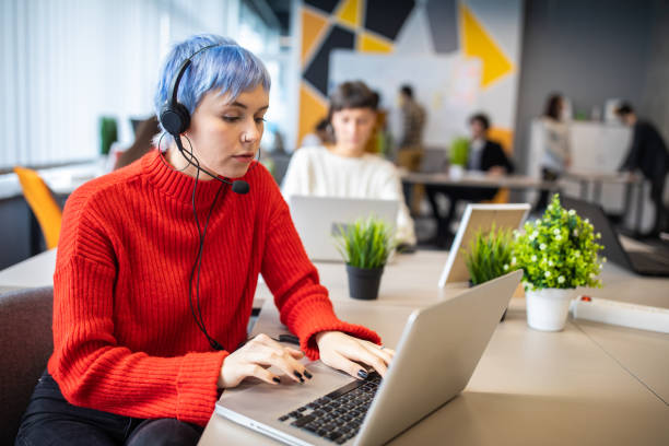 istock Young woman working on laptop with headset in busy modern office 1205741136