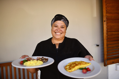A mixed race mid adult female waitress holding two breakfast plates.  She is smiling and wearing black formal restaurant wear.  She is serving two different egg dishes.