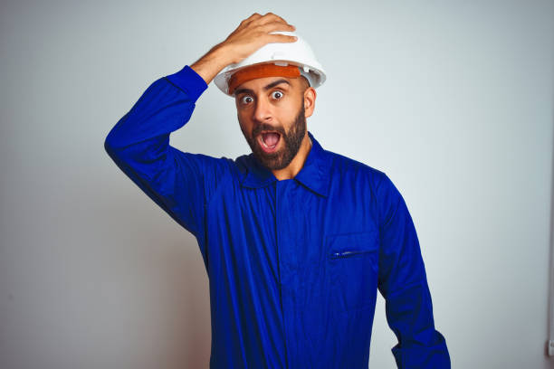 Handsome indian worker man wearing uniform and helmet over isolated white background surprised with hand on head for mistake, remember error. Forgot, bad memory concept.