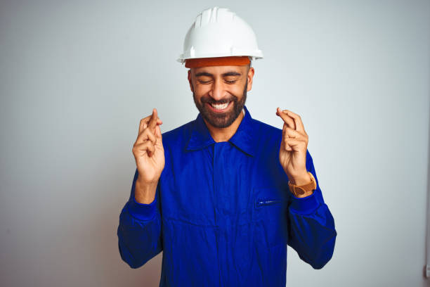 istock Handsome indian worker man wearing uniform and helmet over isolated white background gesturing finger crossed smiling with hope and eyes closed. Luck and superstitious concept. 1205741247