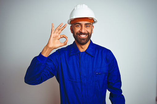 Handsome indian worker man wearing uniform and helmet over isolated white background smiling positive doing ok sign with hand and fingers. Successful expression.