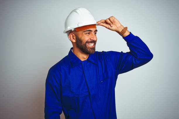 istock Handsome indian worker man wearing uniform and helmet over isolated white background very happy and smiling looking far away with hand over head. Searching concept. 1205741288