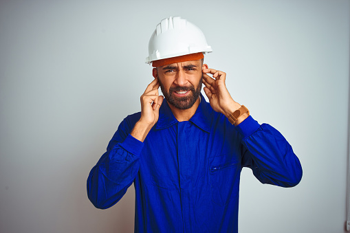 Handsome indian worker man wearing uniform and helmet over isolated white background covering ears with fingers with annoyed expression for the noise of loud music. Deaf concept.