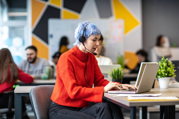 istock Young woman working on laptop with headset in busy modern office 1205741371