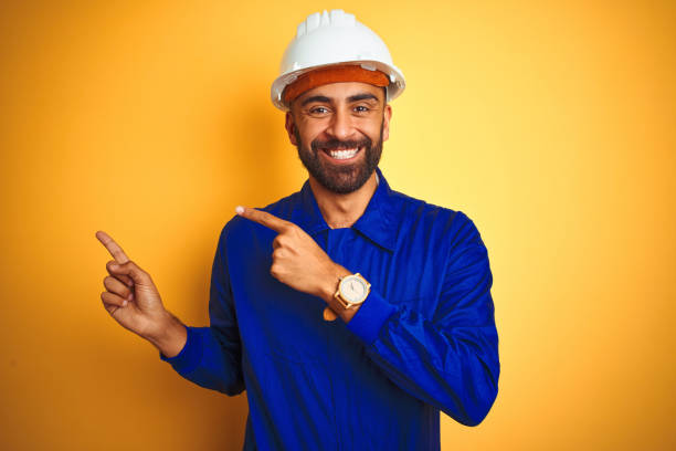 istock Handsome indian worker man wearing uniform and helmet over isolated yellow background smiling and looking at the camera pointing with two hands and fingers to the side. 1205741424
