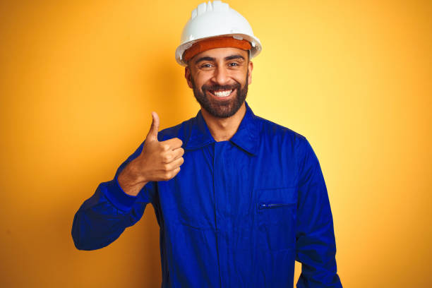 istock Handsome indian worker man wearing uniform and helmet over isolated yellow background doing happy thumbs up gesture with hand. Approving expression looking at the camera with showing success. 1205741452