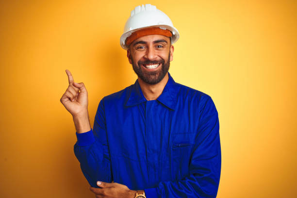 istock Handsome indian worker man wearing uniform and helmet over isolated yellow background with a big smile on face, pointing with hand and finger to the side looking at the camera. 1205741545